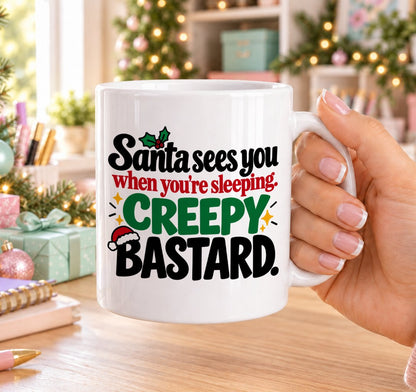 White 11oz ceramic Christmas mug printed with “Santa sees you when you’re sleeping. Creepy bastard.” in red, green and black lettering with Santa hat and holly details, held in a festive room