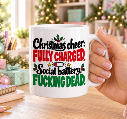 White 11oz ceramic Christmas mug printed with “Christmas cheer: Fully charged. Social battery: F*cking dead.” in red, green and black lettering with a low battery icon, holly and gold stars, held in a festive room