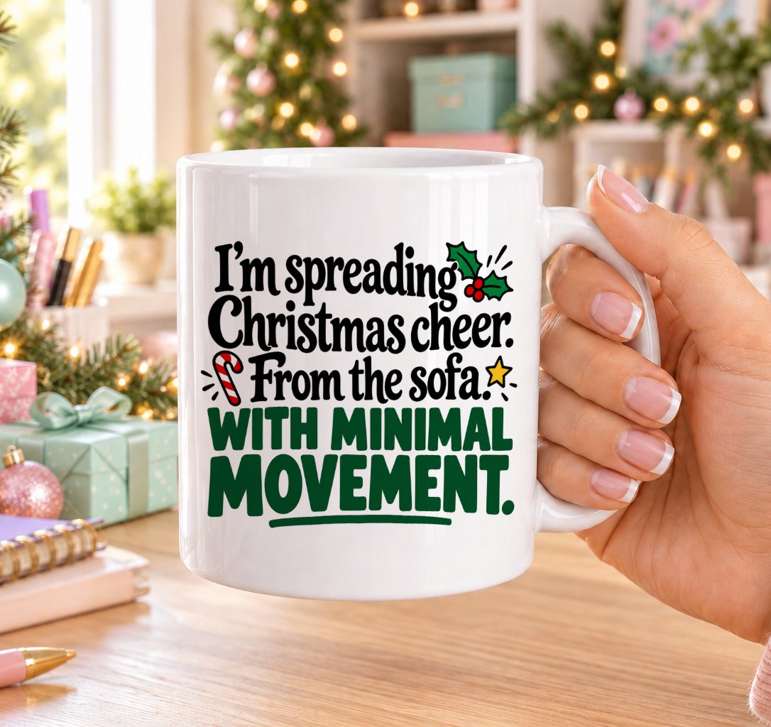 White 11oz ceramic Christmas mug printed with “I’m spreading Christmas cheer. From the sofa. With minimal movement.” in black and green lettering with holly, a candy cane and gold stars, held in a festive room