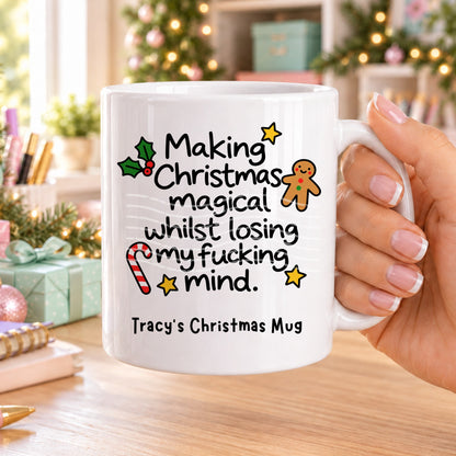 White personalised funny Christmas mug printed with “Making Christmas magical whilst losing my f*cking mind.” with holly, a gingerbread man, candy cane and stars, and the line “Tracy’s Christmas Mug” underneath