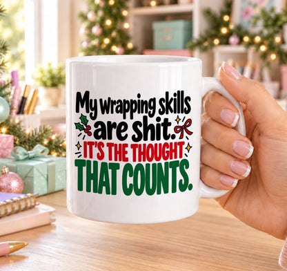 White 11oz ceramic Christmas mug printed with “My wrapping skills are shit. It’s the thought that counts.” in red, green and black lettering with a red bow, holly and gold stars, held in a festive room