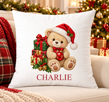 Personalised white Christmas cushion with a teddy in a Santa hat with a pile of presents and a name printed underneath, on a festive sofa