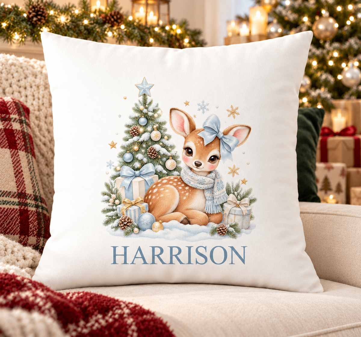 Personalised white Christmas cushion with a fawn with a blue bow beside a frosty Christmas tree and a name printed underneath, on a festive sofa