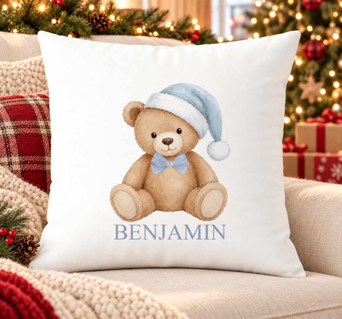 Personalised white Christmas cushion with a teddy bear in a pale blue Santa hat and bow tie and a name printed underneath, on a festive sofa