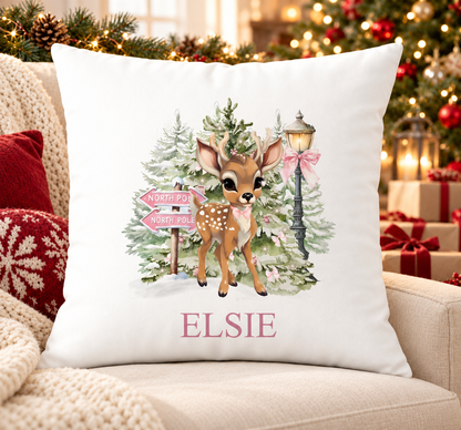 Personalised white Christmas cushion with a fawn beside a pink North Pole signpost in a snowy forest and a name printed underneath, on a festive sofa