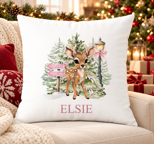 Personalised white Christmas cushion with a fawn beside a pink North Pole signpost in a snowy forest and a name printed underneath, on a festive sofa