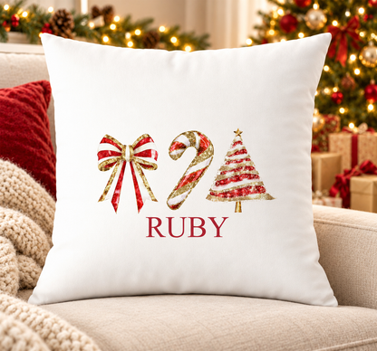 Personalised white Christmas cushion with red bow, candy cane and Christmas tree trio in red and gold and a name printed underneath, on a festive sofa