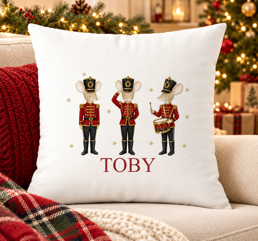 Personalised white Christmas cushion with three mice dressed as nutcracker soldiers and a name printed underneath, on a festive sofa