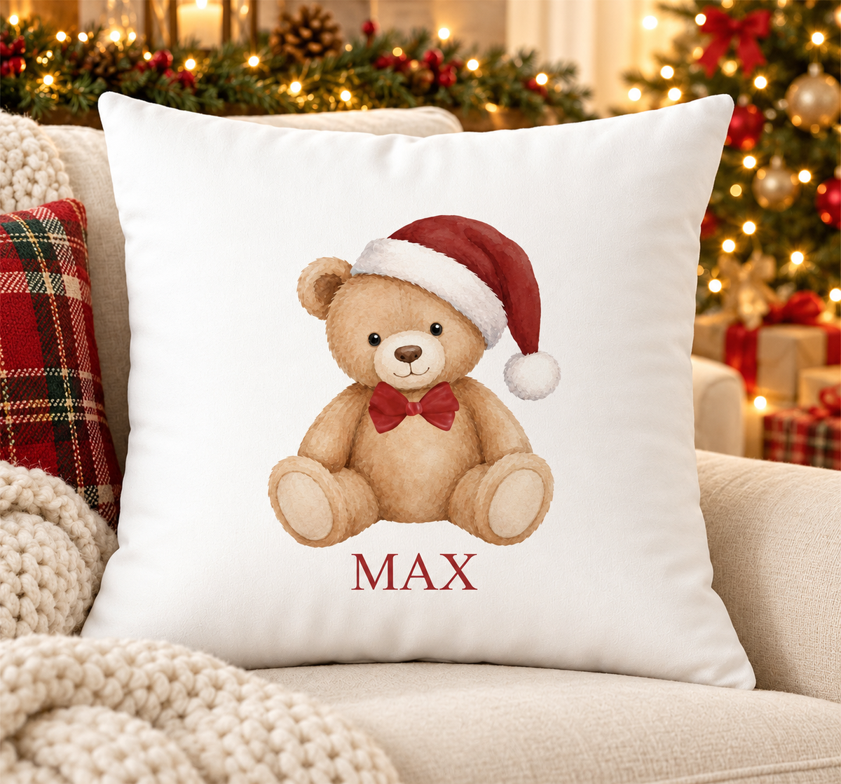 Personalised white Christmas cushion with a teddy bear in a red Santa hat and bow tie and a name printed underneath, on a festive sofa