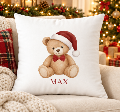 Personalised white Christmas cushion with a teddy bear in a red Santa hat and bow tie and a name printed underneath, on a festive sofa