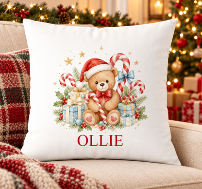 Personalised white Christmas cushion with a teddy in a Santa hat with candy canes and presents and a name printed underneath, on a festive sofa