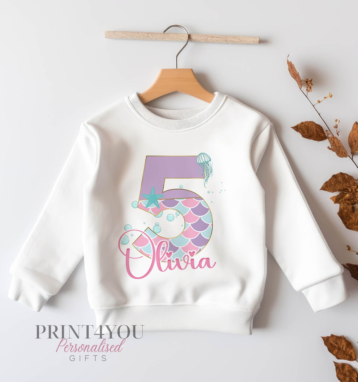 White children's sweatshirt on a hanger printed with a pastel mermaid tail number 5, jellyfish, starfish and the name Olivia